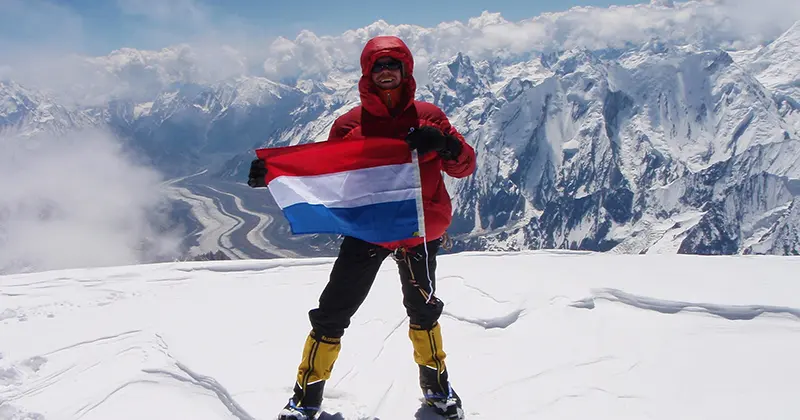 Een persoon in rode winterkleding staat op een besneeuwde bergtop met een rood-wit-blauwe vlag in de hand; op de achtergrond zijn met sneeuw bedekte bergen en een kronkelende rivier te zien.